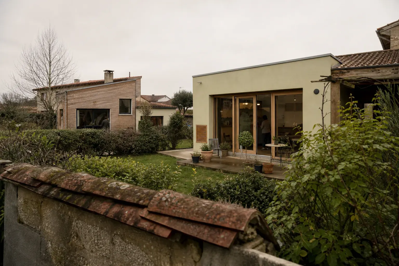 Deux maisons voisines séparées par une clôture basse, l’une avec une extension maçonnée, l’autre en bois